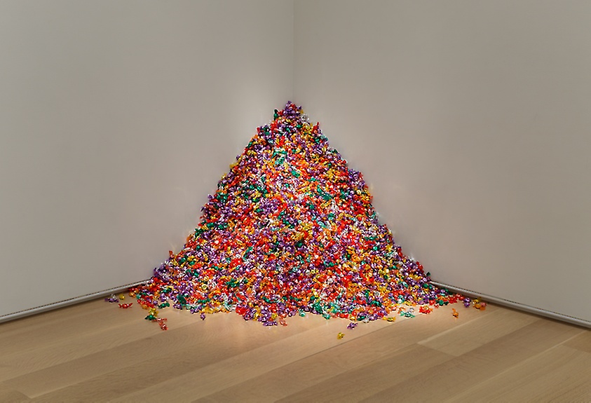 175 pounds of hard candy wrapped in green, purple, orange, gold, and other colored foil is piled in the corner of a white-walled museum gallery, spilling across the floor