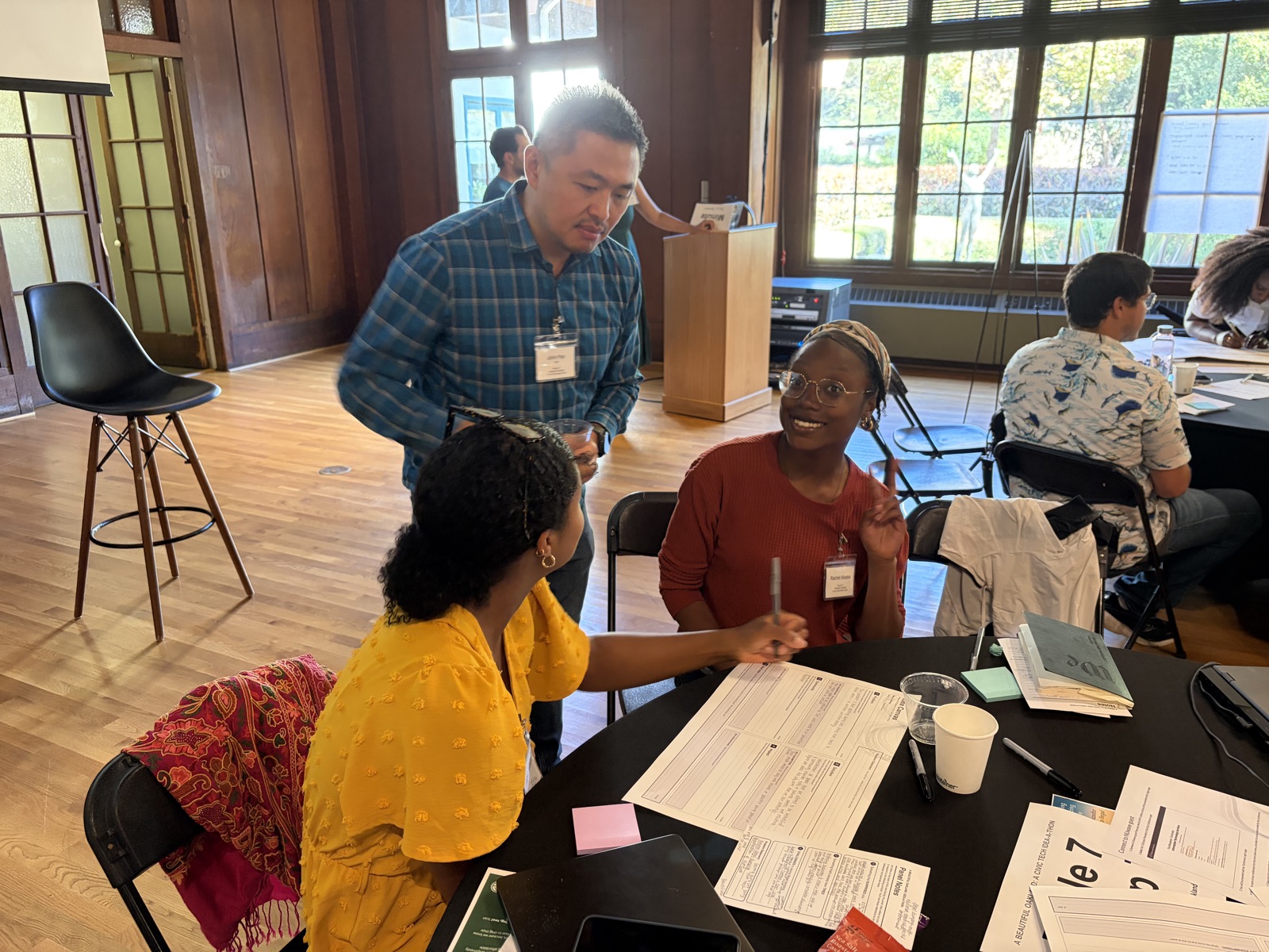 Idea-A-Thon attendees talking around a table covered in worksheets and Panel Notes cards, with other groups working in the background
